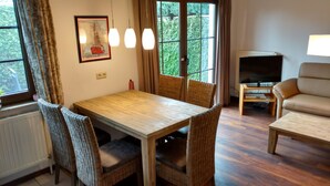 A dining area with a wooden table and wicker chairs, a television on a wooden stand, and a sofa in a living room. - Holiday house, WiFi, close to the beach, max. 6 people, 80sqm, 3 bedrooms, barbecue area  (Butjadingen)