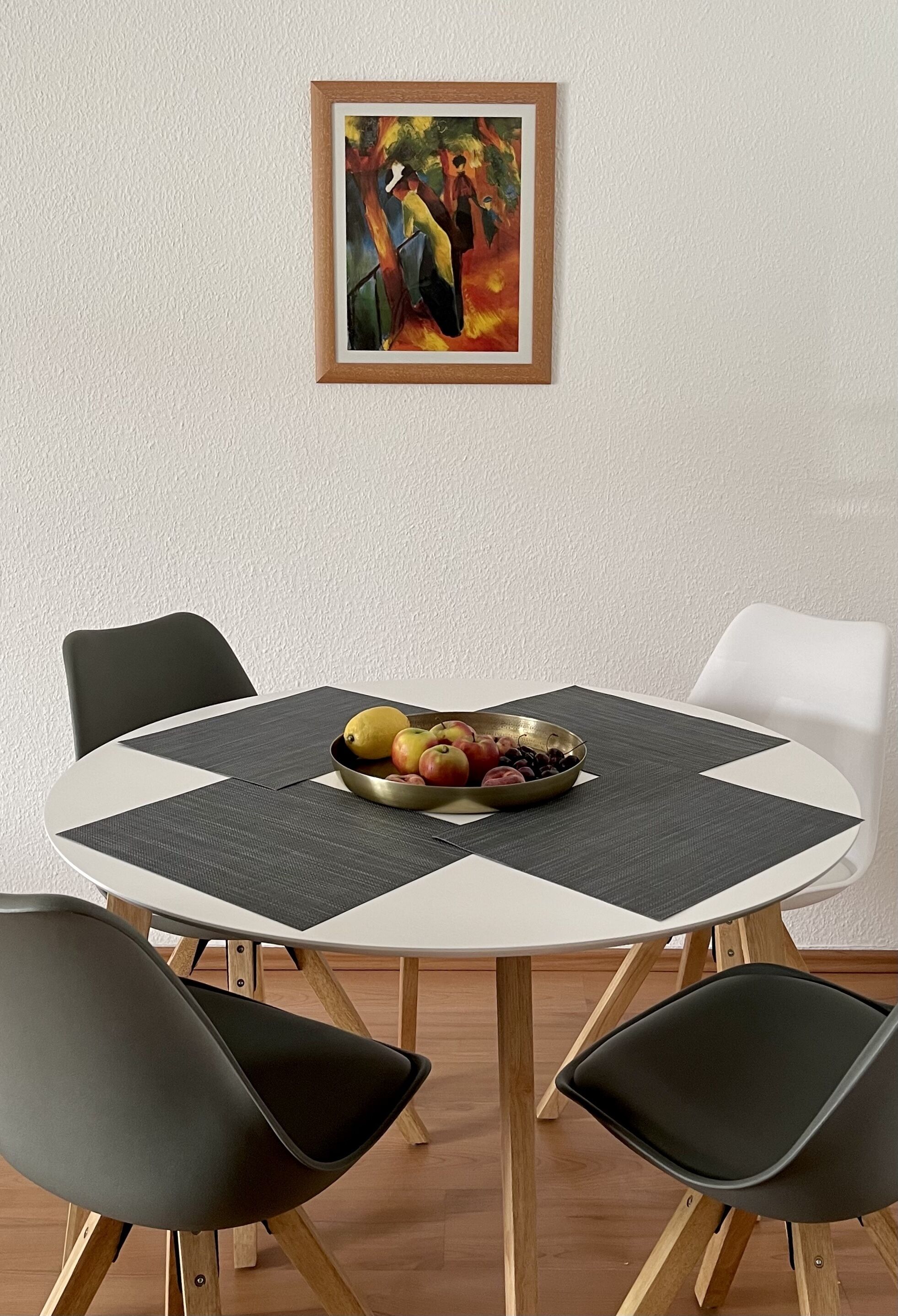A round dining table with a white and black placemat, surrounded by four black chairs. A bowl of fruit is placed on the table.