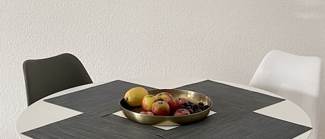 A round dining table with a white and black placemat, surrounded by four black chairs. A bowl of fruit is placed on the table.