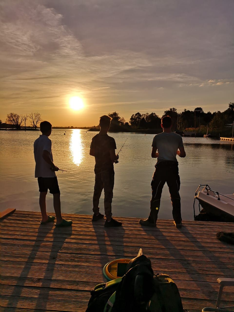 Three people fishing on a wooden dock at sunset.