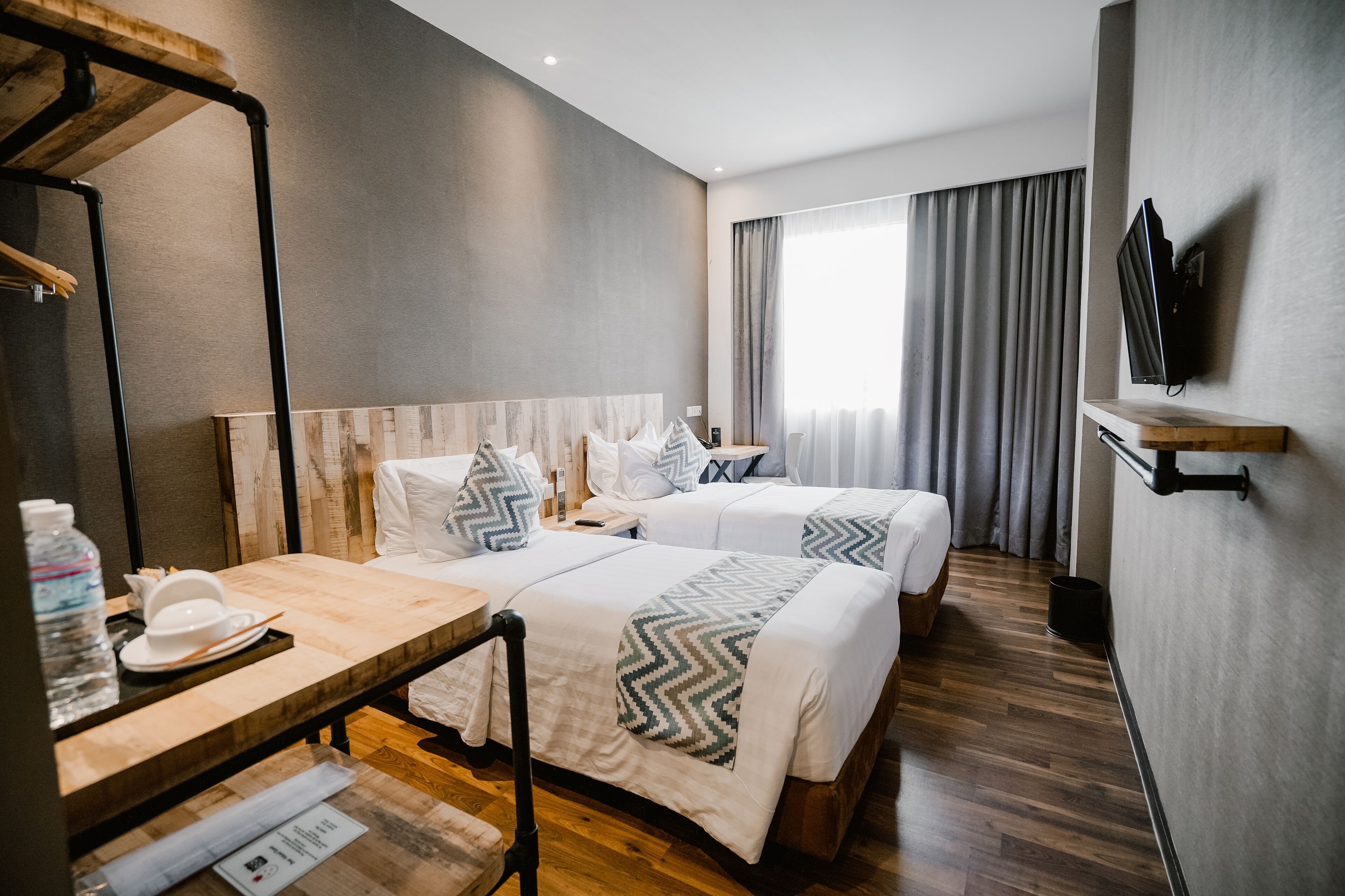 Habitación de hotel con dos camas, una mesa de madera con una botella de agua, un televisor en la pared y un ventanal con cortinas.