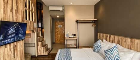 A modern hotel room with a wooden accent wall, a large bed with patterned pillows, a flat-screen TV mounted on the wall, and a small table with a coffee maker.