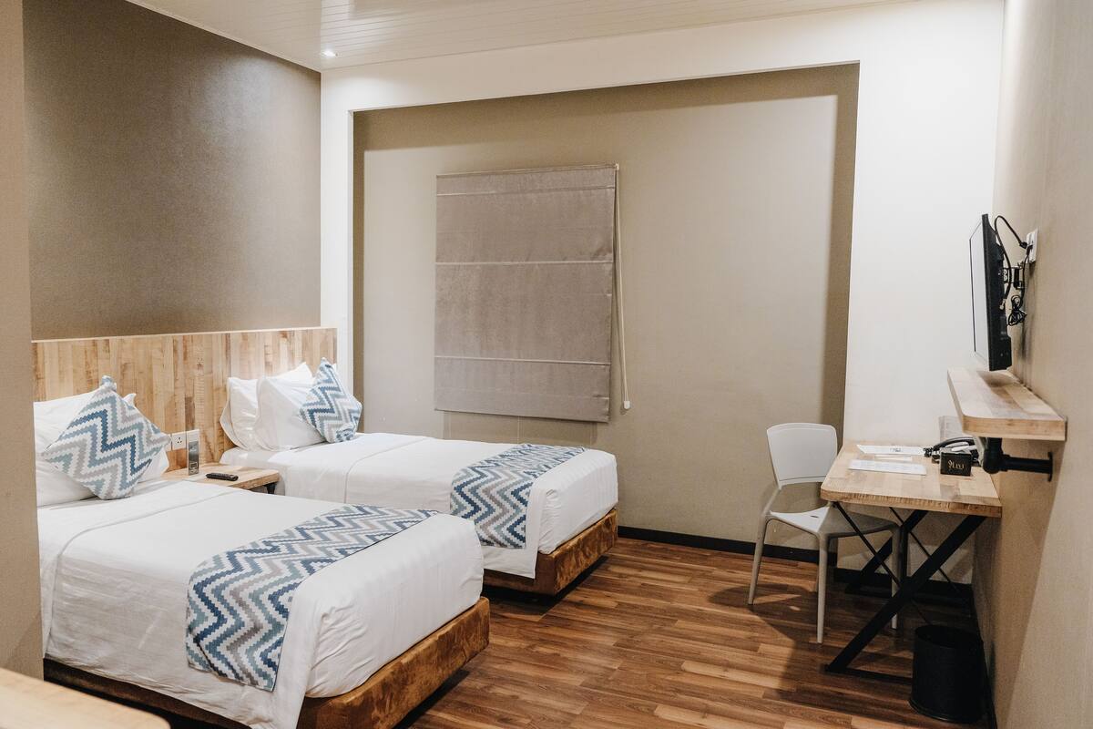 Une chambre d’hôtel avec deux lits, un bureau, une chaise, un plancher en bois et une fenêtre avec un rideau.