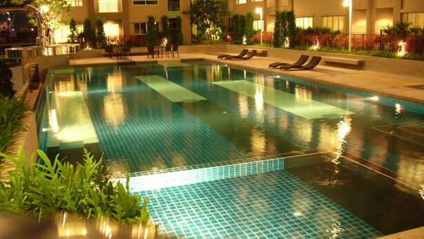 Una piscina con azulejos azules cristalinos, rodeada de sillas de descanso y un edificio con varios balcones.