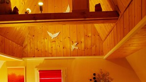 Ein gemütliches Schlafzimmer mit Holzbalkendecke, einem Bett mit rot-weißer Bettwäsche, zwei Nachttischen mit Lampen und einem Fenster mit roten Jalousien.