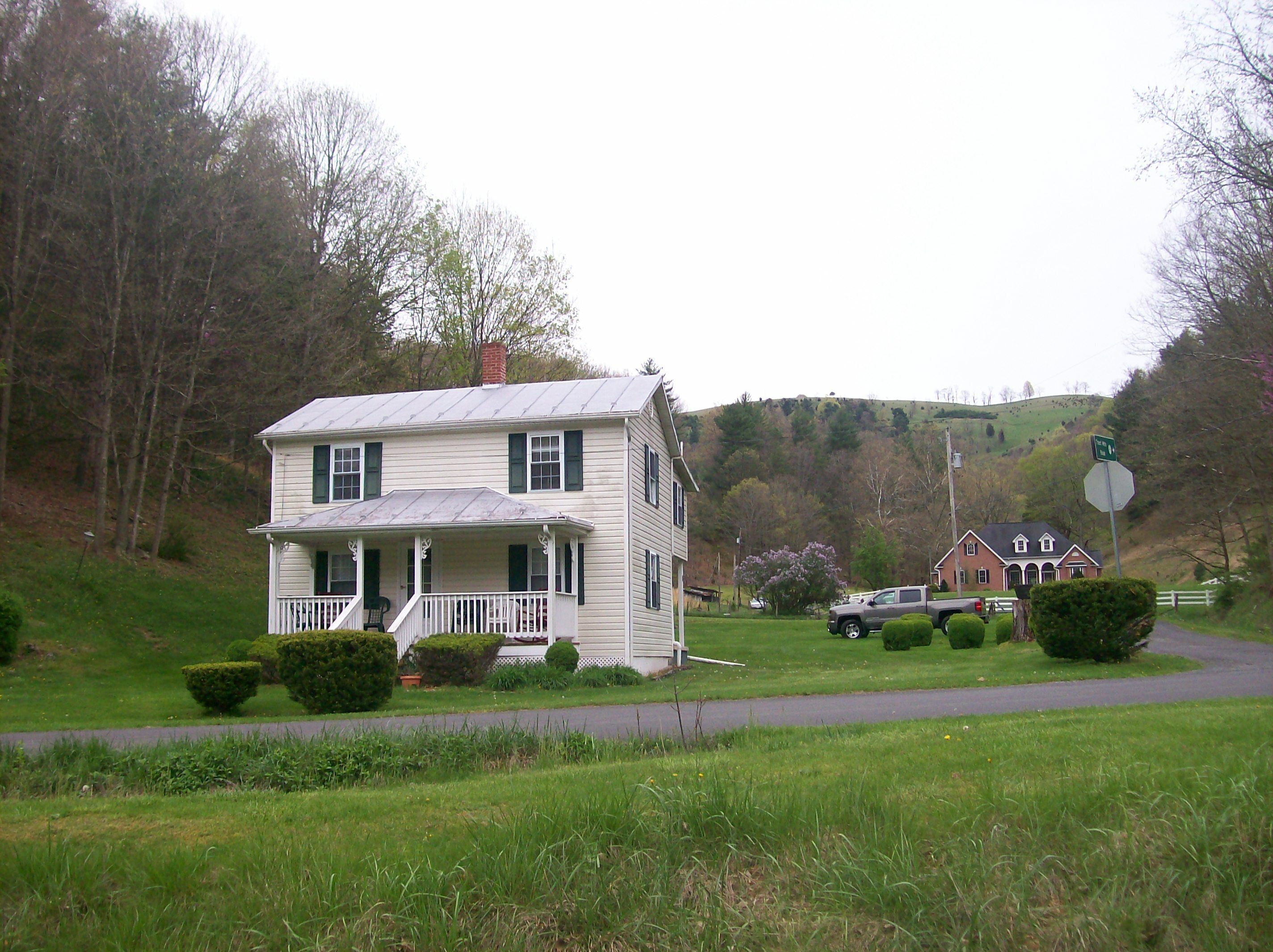A two-story house with a porch, surrounded by greenery and a hill in the background.