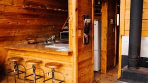 A wooden cabin interior with a kitchen area, bar stools, a refrigerator, and a wood-burning stove.