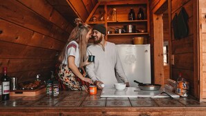 A cozy wooden cabin kitchen with a refrigerator, shelves with bottles and jars, a coffee maker, and a countertop with a bowl and a frying pan.