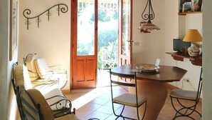 Ein gemütliches Zimmer mit Holzbalkendecke, einem Esstisch mit Stühlen, einer Couch und einer kleinen Küchenzeile mit Lampe und Regal.