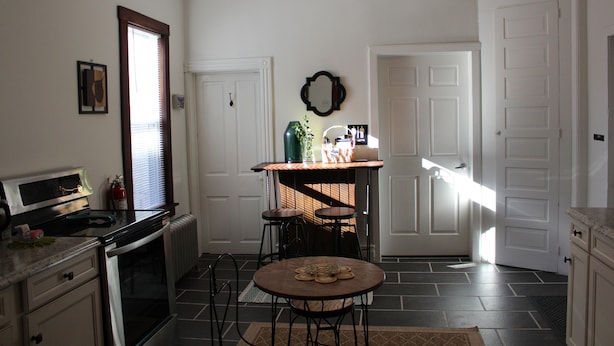 Una cocina con estufa, horno y un comedor con taburetes. Hay una ventana con persianas, una puerta y un espejo montado en la pared.