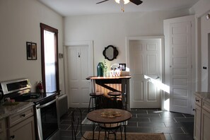 A kitchen with a stove, oven, and a dining area with stools. There is a window with blinds, a door, and a wall-mounted mirror. - Glen Onoko Getaway - Antique Transoms, 10-Foot Ceilings & New King Bed (Jim Thorpe)