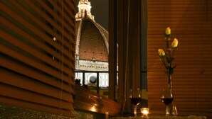 Una habitación con vistas a un edificio histórico, un florero con flores y dos copas de vino tinto sobre una mesa.