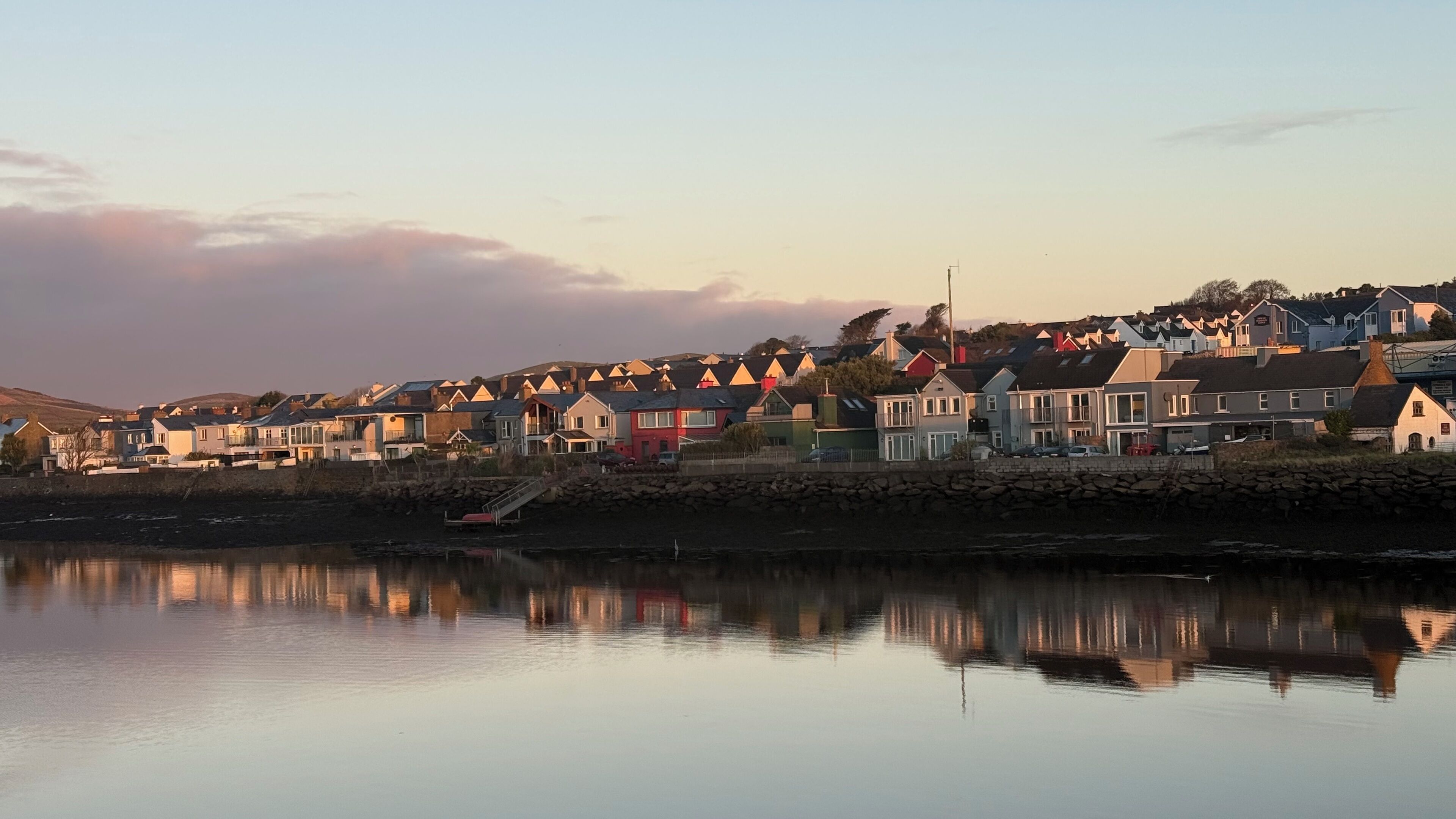 A coastal residential area with houses along the water's edge, a calm lake reflecting the buildings, and a clear sky.