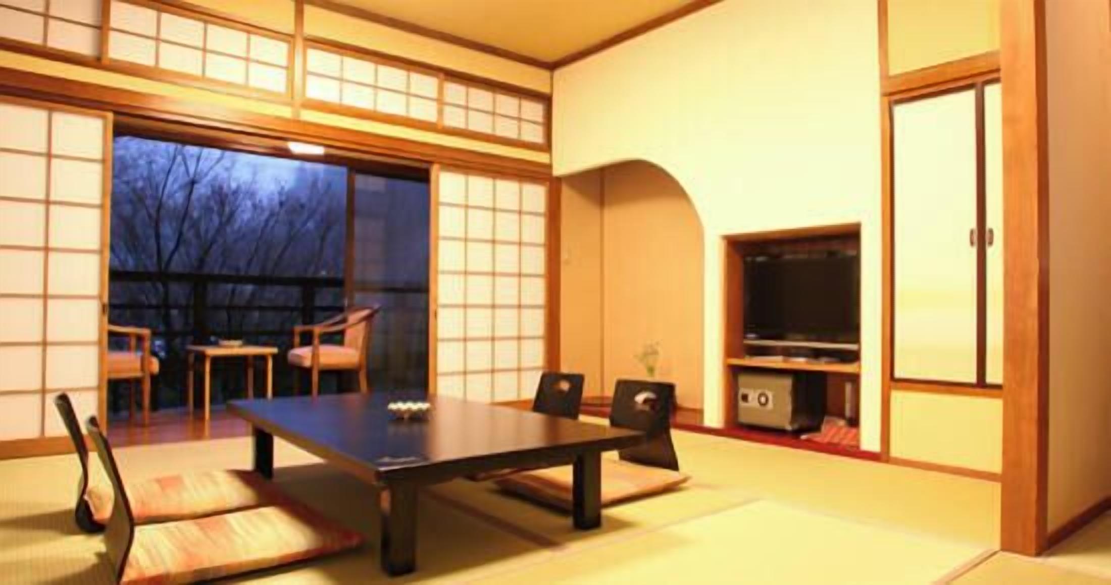 Ein traditioneller japanischer Raum mit Tatami-Boden, einem niedrigen Tisch, Stühlen, einem Fernseher und Schiebetüren.