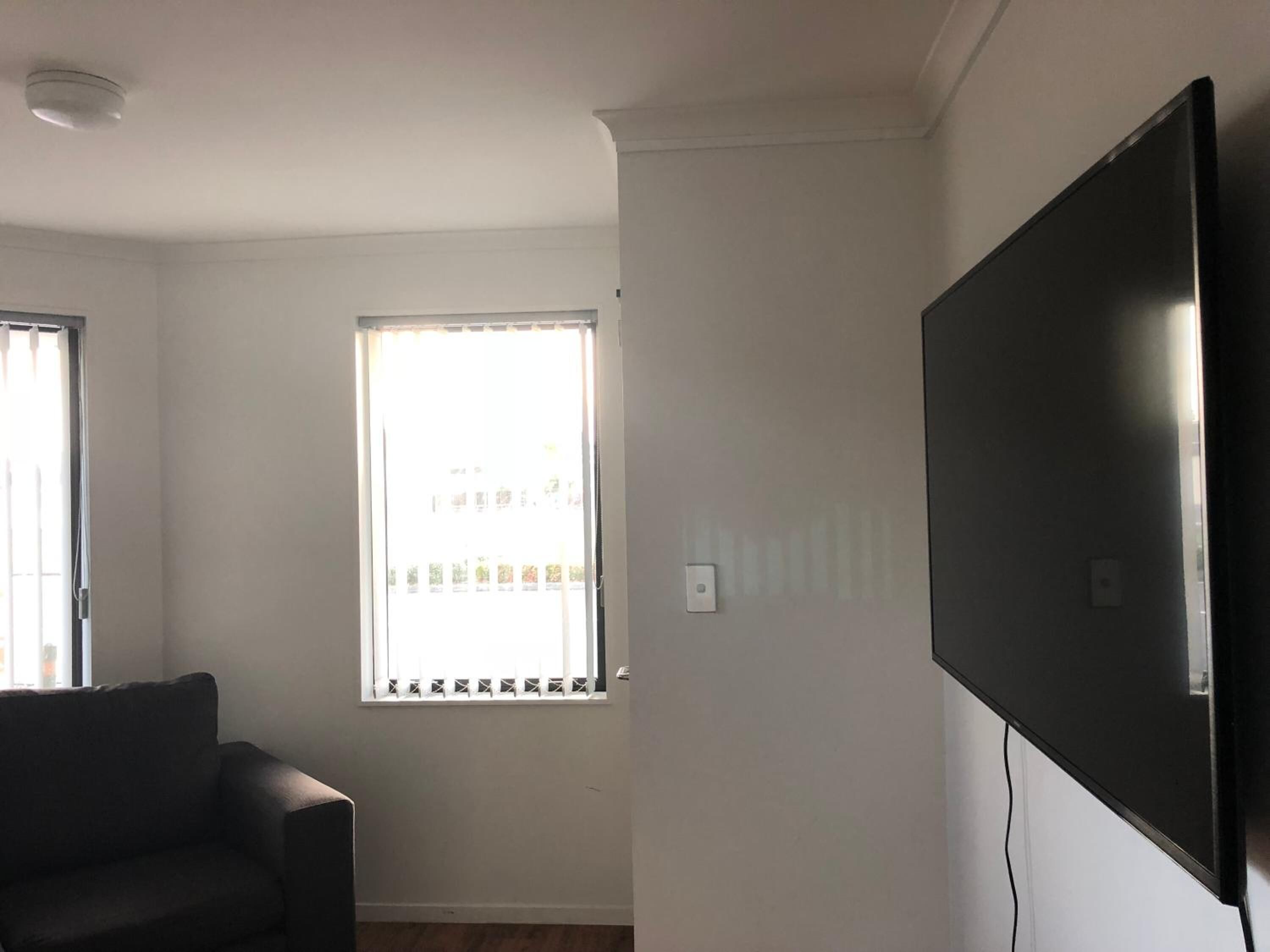 A room with a large flat-screen TV mounted on the wall, a window with blinds, and a dark-colored sofa.