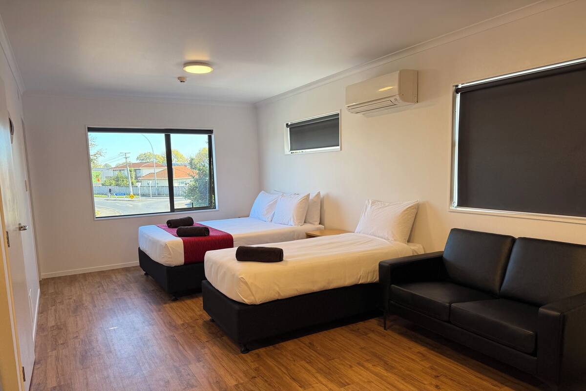 Habitación de hotel con dos camas, un sofá negro, una ventana con vista a la calle y un equipo de aire acondicionado.