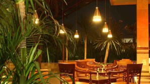 An outdoor dining area with wooden tables and chairs, surrounded by lush greenery and palm trees. - Bali World Hotel (Bandung)