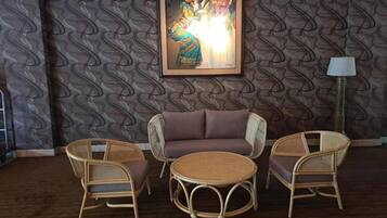 Une pièce avec un papier peint à motifs, un canapé, deux fauteuils en osier, une table ronde et une œuvre d’art encadrée accrochée au mur.