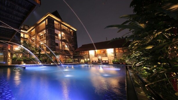A nighttime view of a hotel pool area with a water feature, surrounded by lush greenery and a multi-story building with a warm-lit facade. - Bali World Hotel (Bandung)
