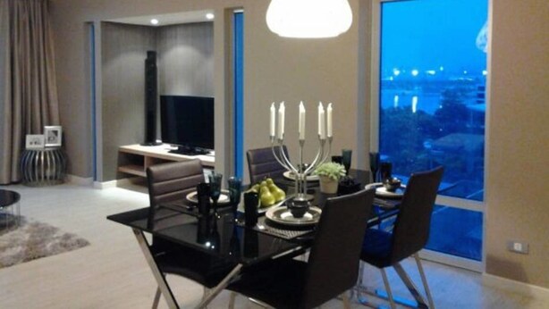 A modern living room with a dining table set for four, a large window with a city view, and a television in the background. - The Lake 3 Condominium (Pak Kret)