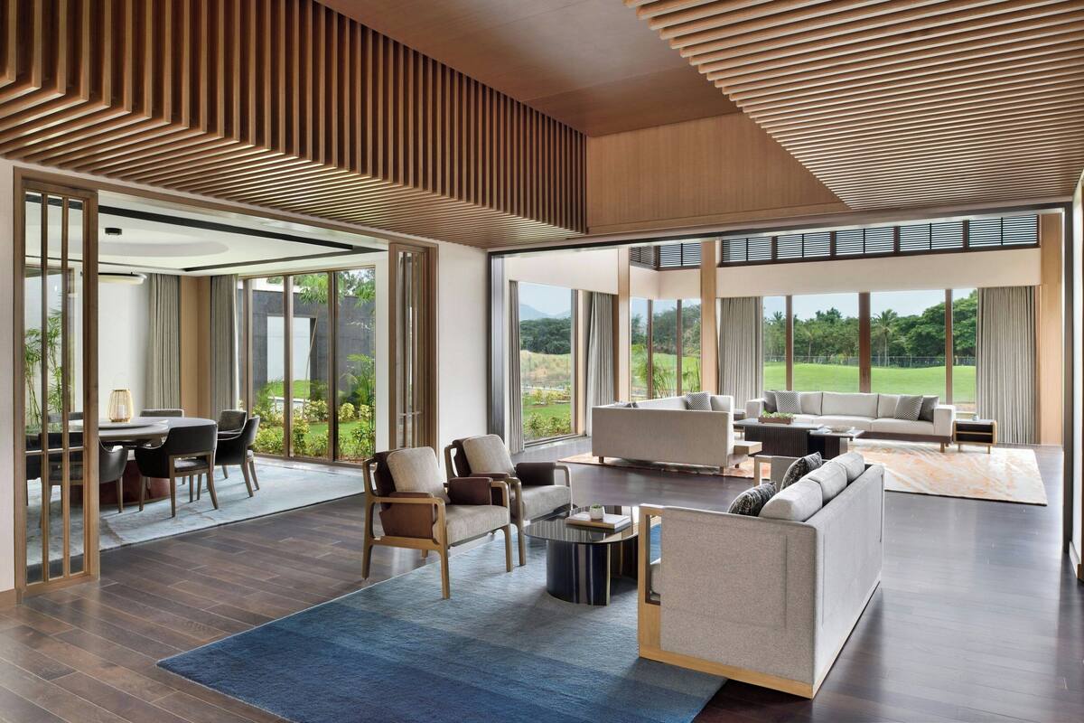 A spacious living area with a large sofa, armchairs, and a coffee table. There is a dining area with a table and chairs. The room has large windows offering a view of the outdoors.