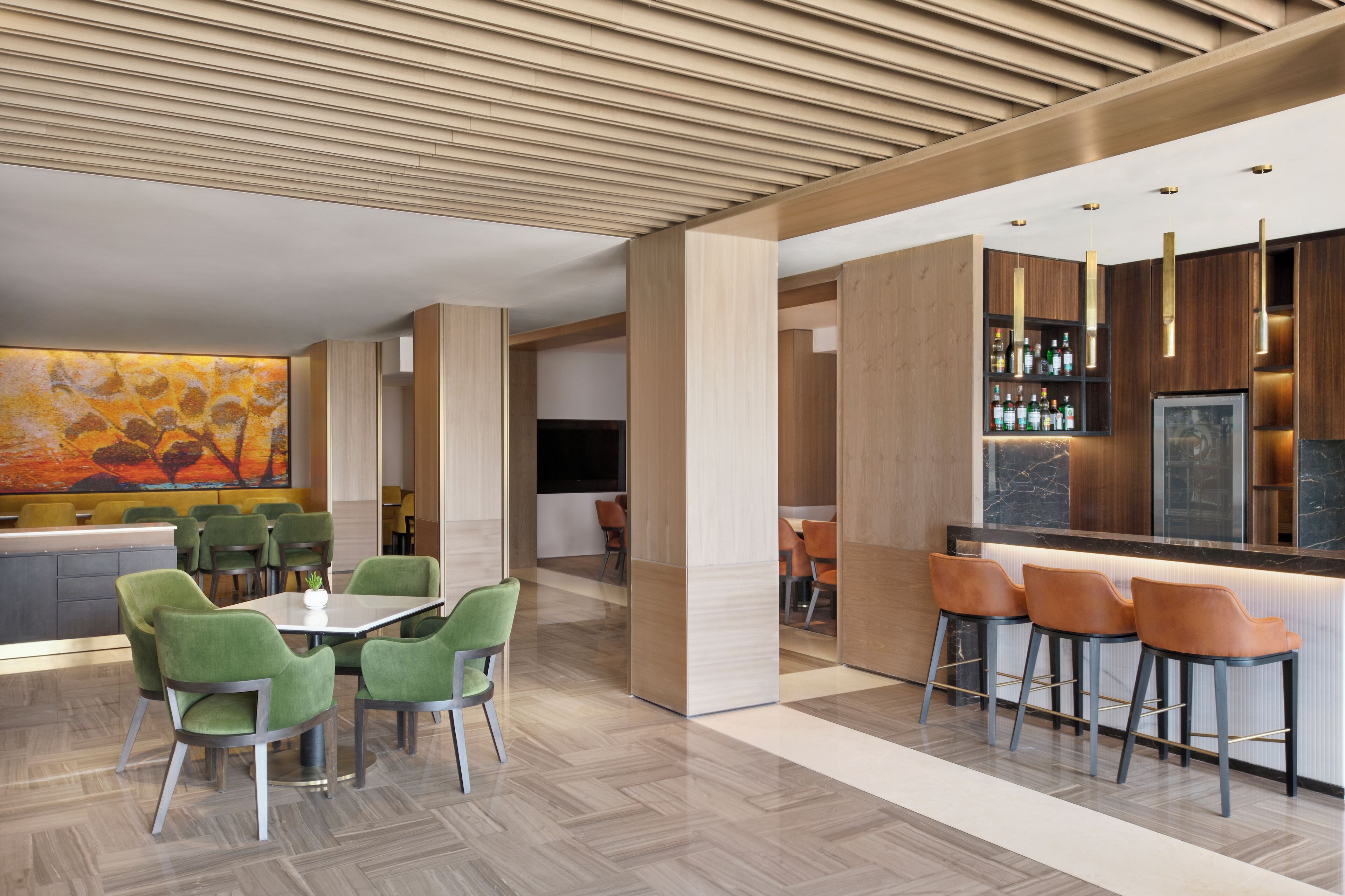 A modern bar area with a wooden bar counter, green and orange bar stools, and a large abstract painting on the wall.