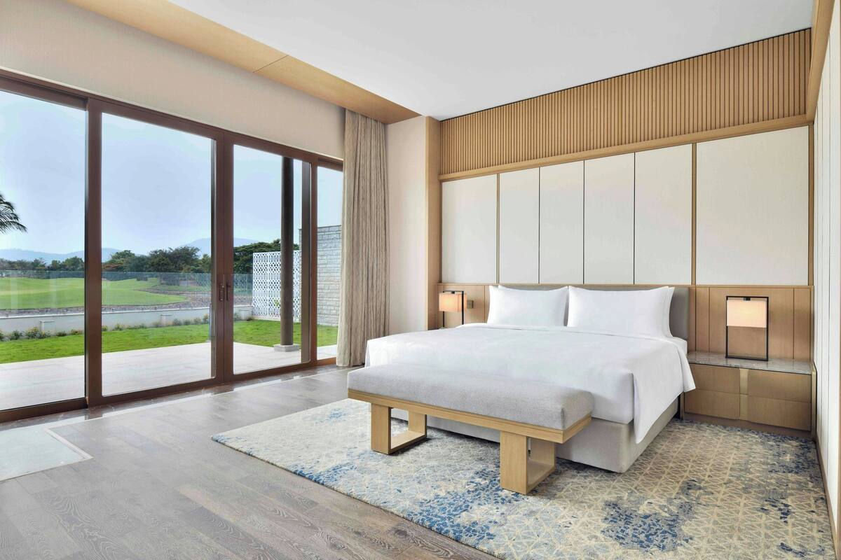 A modern hotel room with a large bed, a wooden headboard, and a view of a golf course through floor-to-ceiling windows.