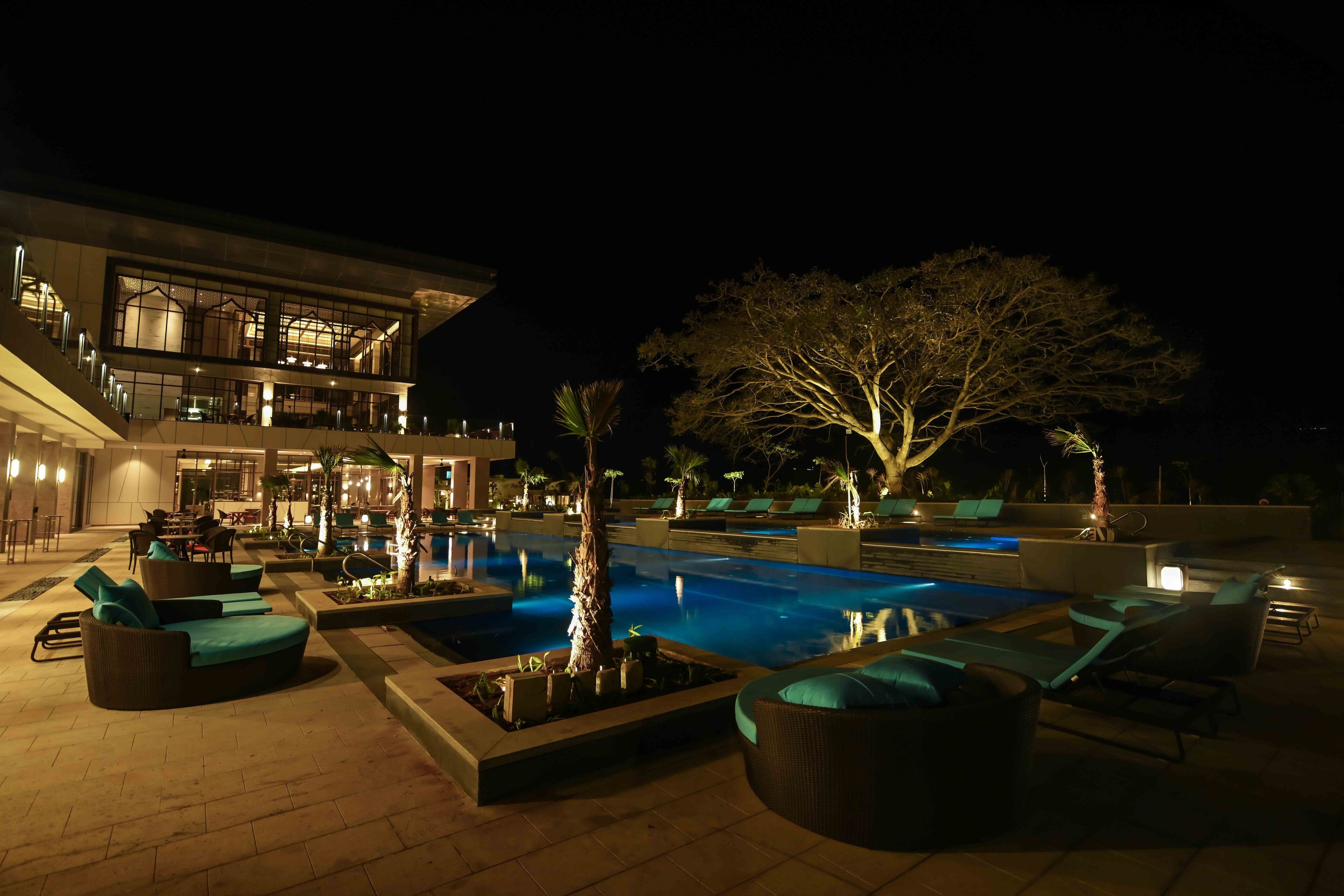 A nighttime pool area with lounge chairs and a large tree.