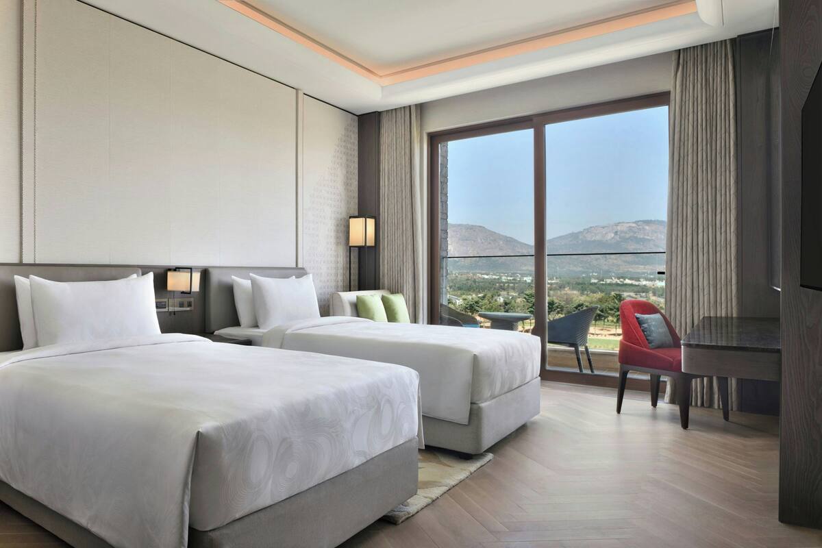 A hotel room with two beds, a desk, a chair, and a large window offering a view of the outdoors.