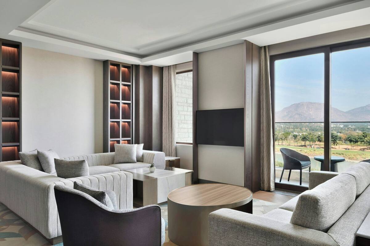 A modern hotel room with a large window offering a mountain view, a sofa, a coffee table, and a TV.