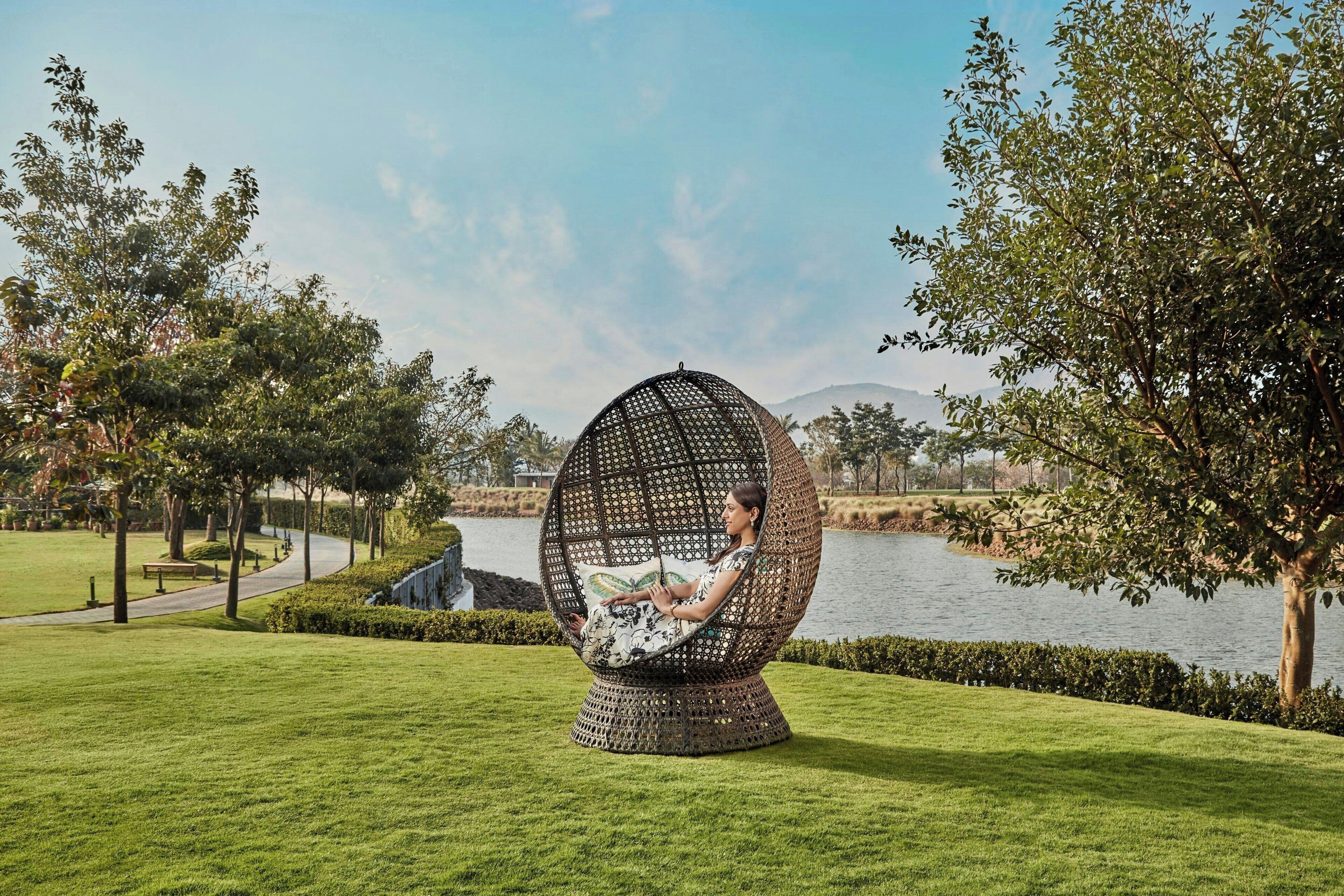A person sitting in a large, spherical, woven chair with a cushion, in a park with a lake and trees.