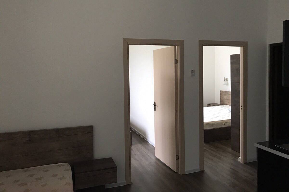 Un dormitori amb un panell de fusta al capçal del llit, un llit i una cambrera de nit. Hi ha dues portes que donen a un altre espai.