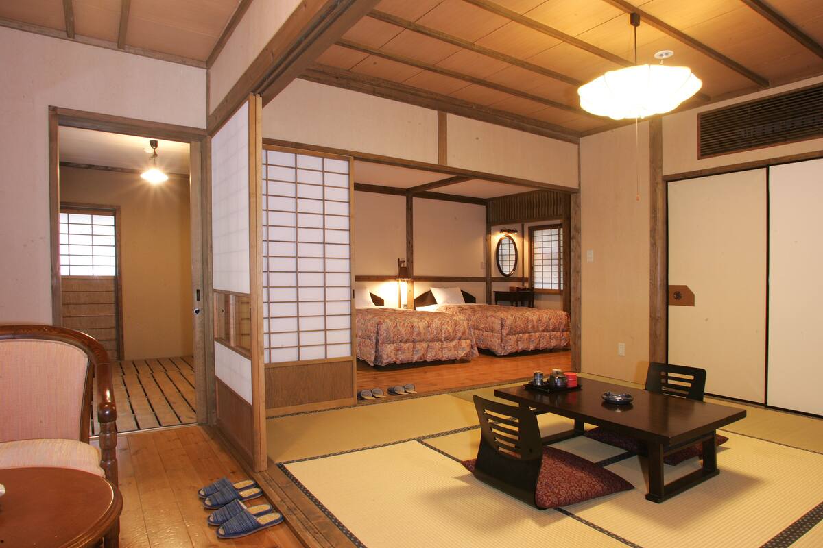A traditional Japanese room with tatami flooring, a low table with chairs, and a bed with a patterned bedspread.