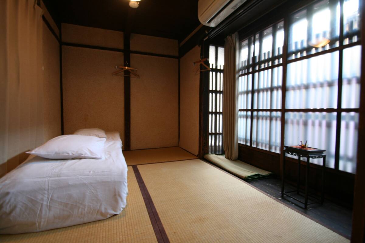 Une pièce de style japonais traditionnel, avec un sol recouvert de tatamis, un futon servant de lit, une porte coulissante et une petite table avec une lampe.