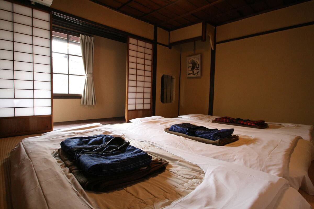 Une pièce de style japonais traditionnel, avec un sol recouvert de tatamis, deux lits où les vêtements sont soigneusement pliés, et des paravents en papier de riz (shoji).