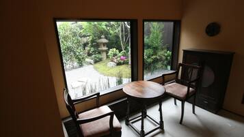 Quarto com uma grande janela que oferece vista para um jardim com um candeeiro de pedra e flores cor-de-rosa. Há uma mesa de madeira redonda com duas cadeiras.