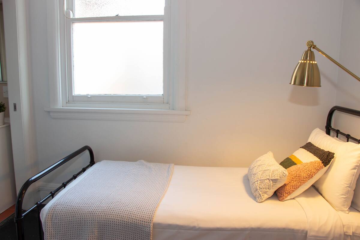 A small, neatly made bed with a white blanket and pillows, a bedside lamp, and a window with blinds.