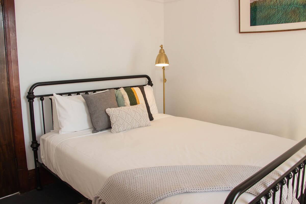A neatly made bed with a white comforter and multiple pillows, a gold floor lamp, and a wooden door to the left.