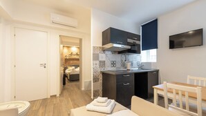 A compact hotel room with a kitchenette, a bed with a neatly folded towel, a small dining table, and a TV mounted on the wall. - Flaminia Rooms&Suites (Rome)
