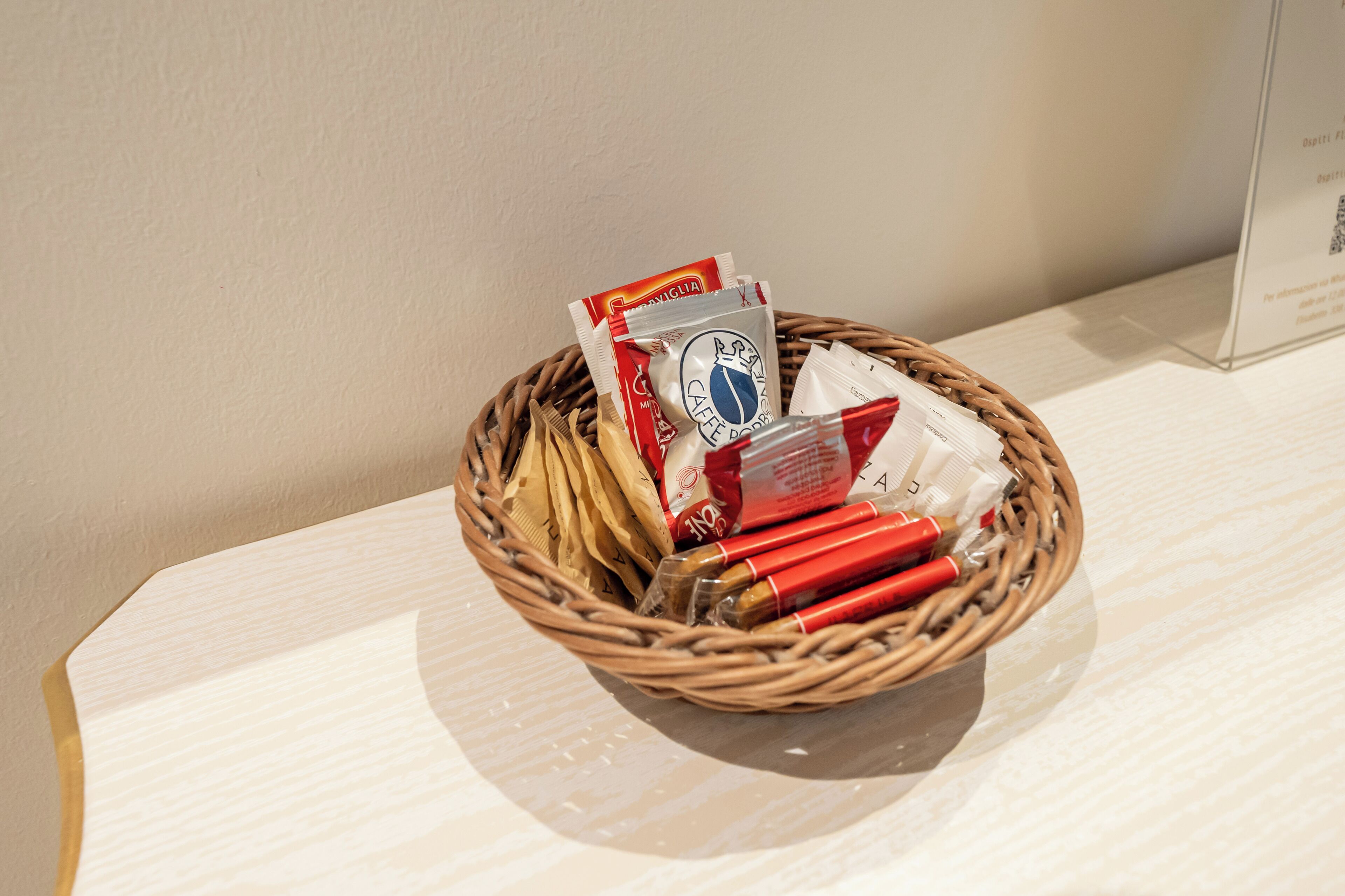 Una cesta de mimbre con varios objetos, incluyendo un paquete rojo y blanco, una bolsa de plástico transparente y algunos objetos cilíndricos rojos.