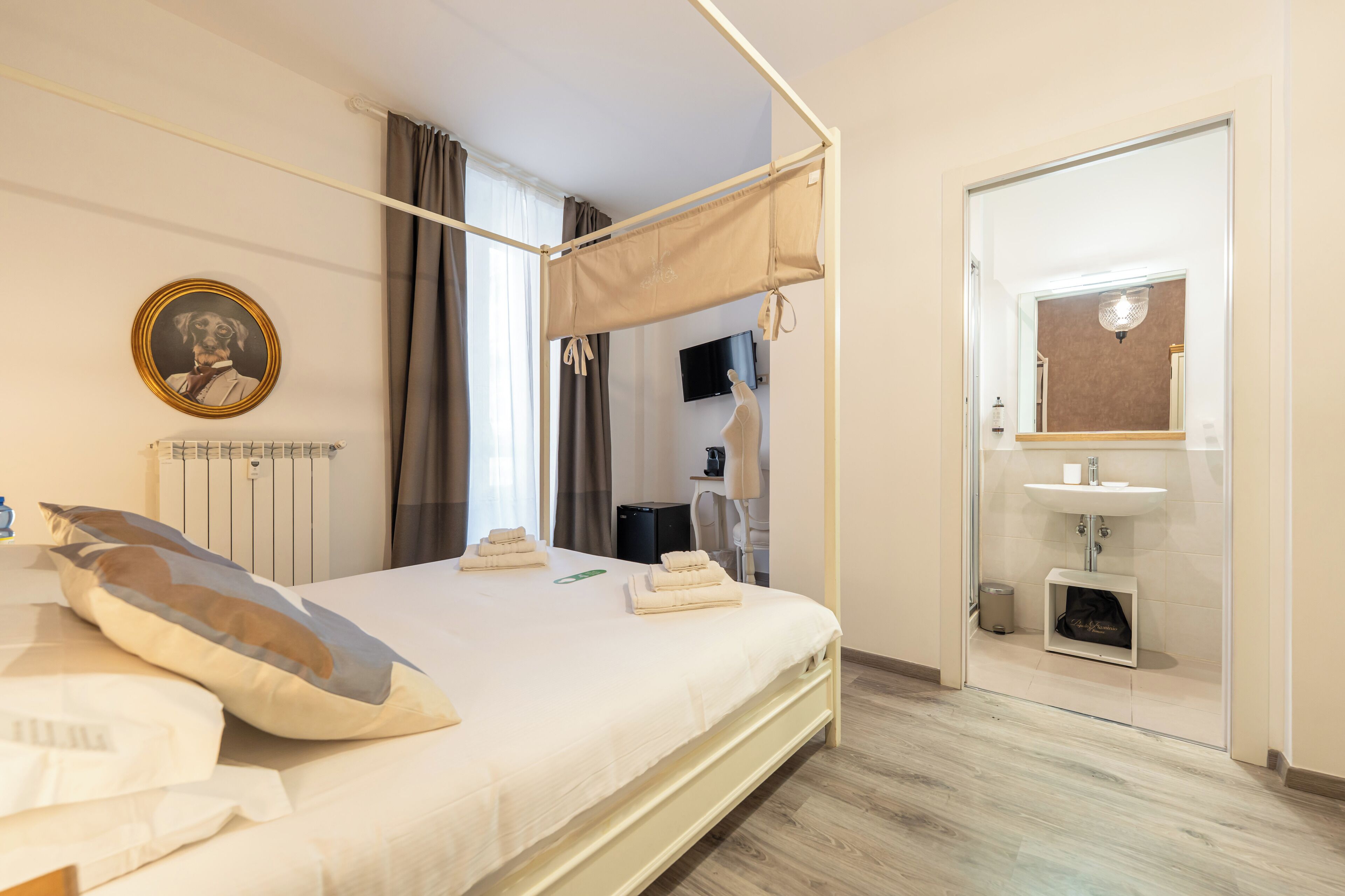 A neatly made bed with a canopy, a bedside table, a lamp, a framed picture on the wall, and a bathroom with a sink and mirror.