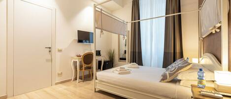 A hotel room with a four-poster bed, a desk with a chair, a TV, and a bedside table with a bottle and remote control.