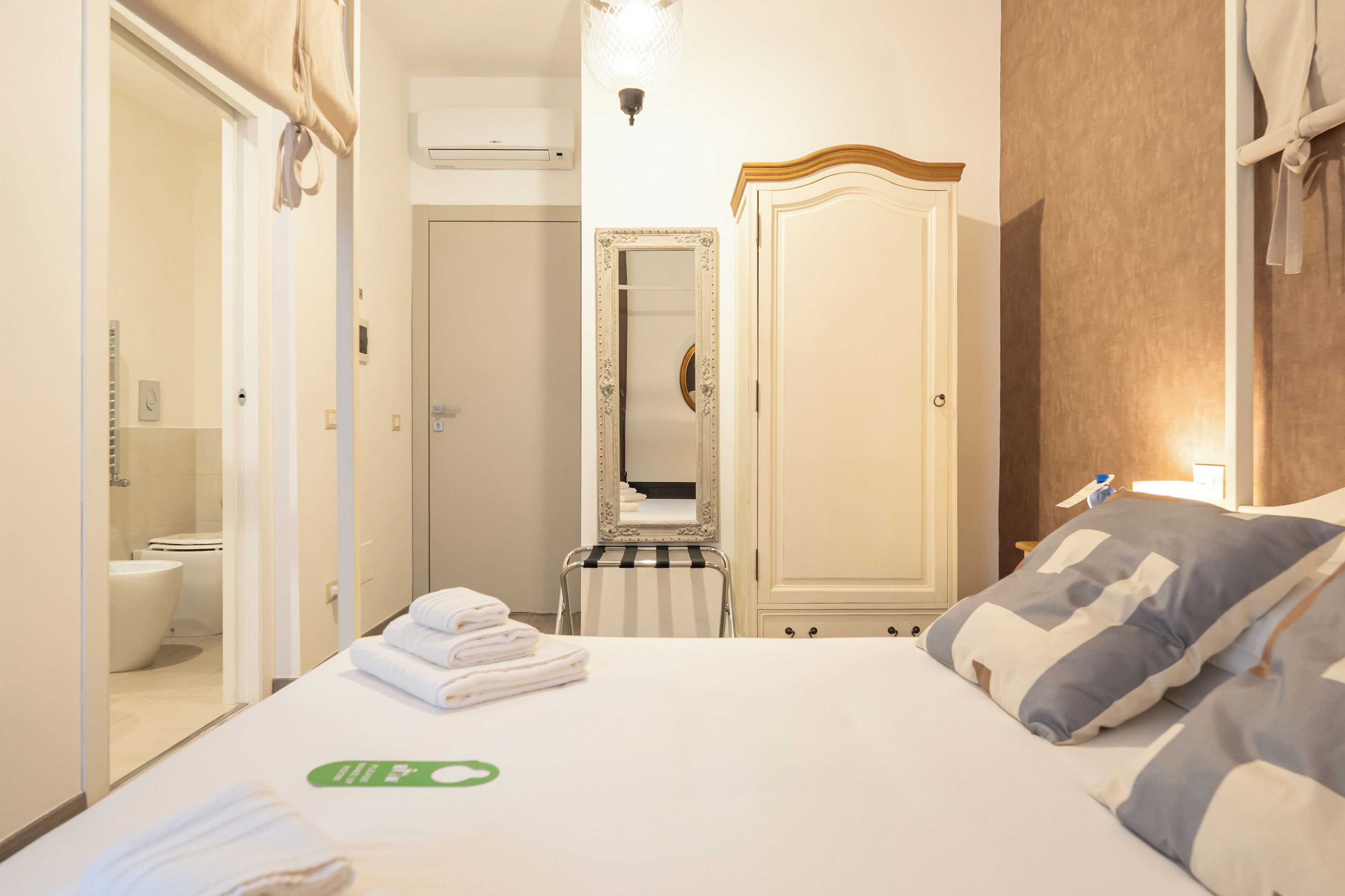 Habitación de hotel con cama, mesita de noche, espejo, armario y baño visible a través de una puerta abierta.