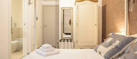 Habitación de hotel con cama, mesita de noche, espejo, armario y baño visible a través de una puerta abierta.