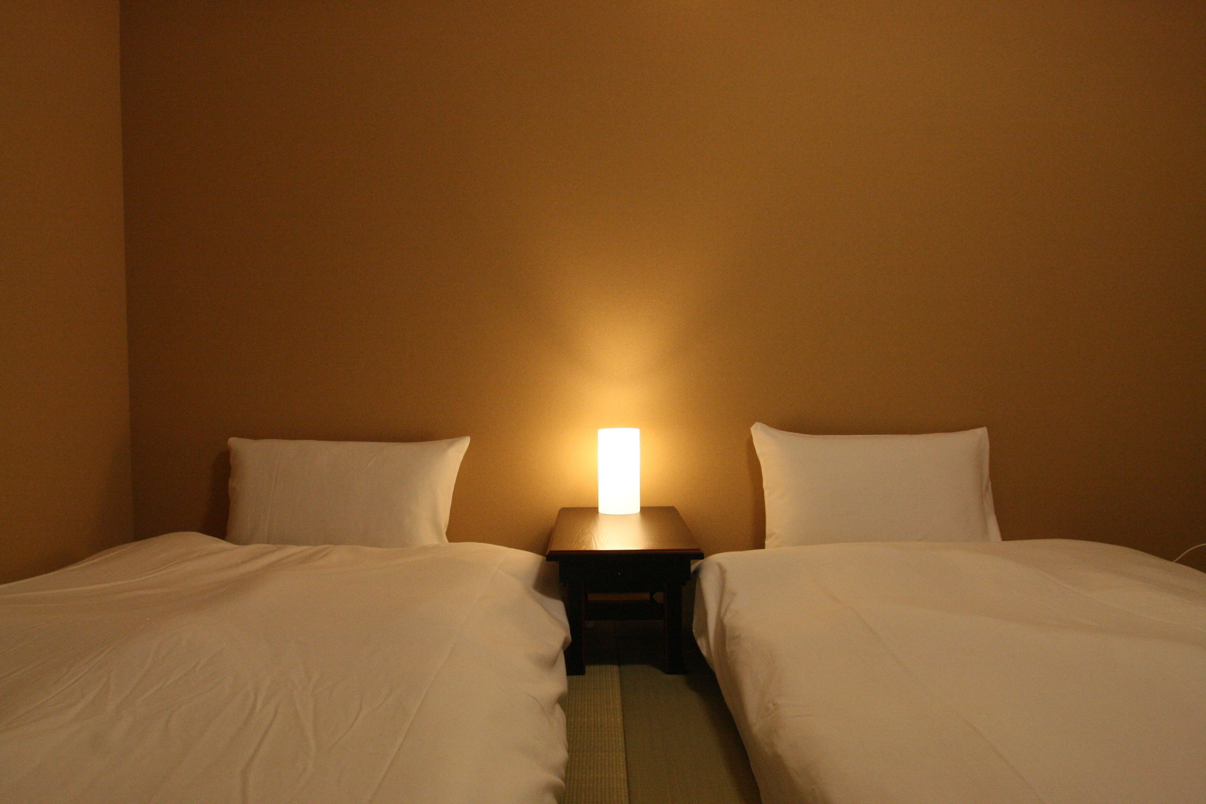 A hotel room with two beds, a nightstand with a lamp, and plain walls.