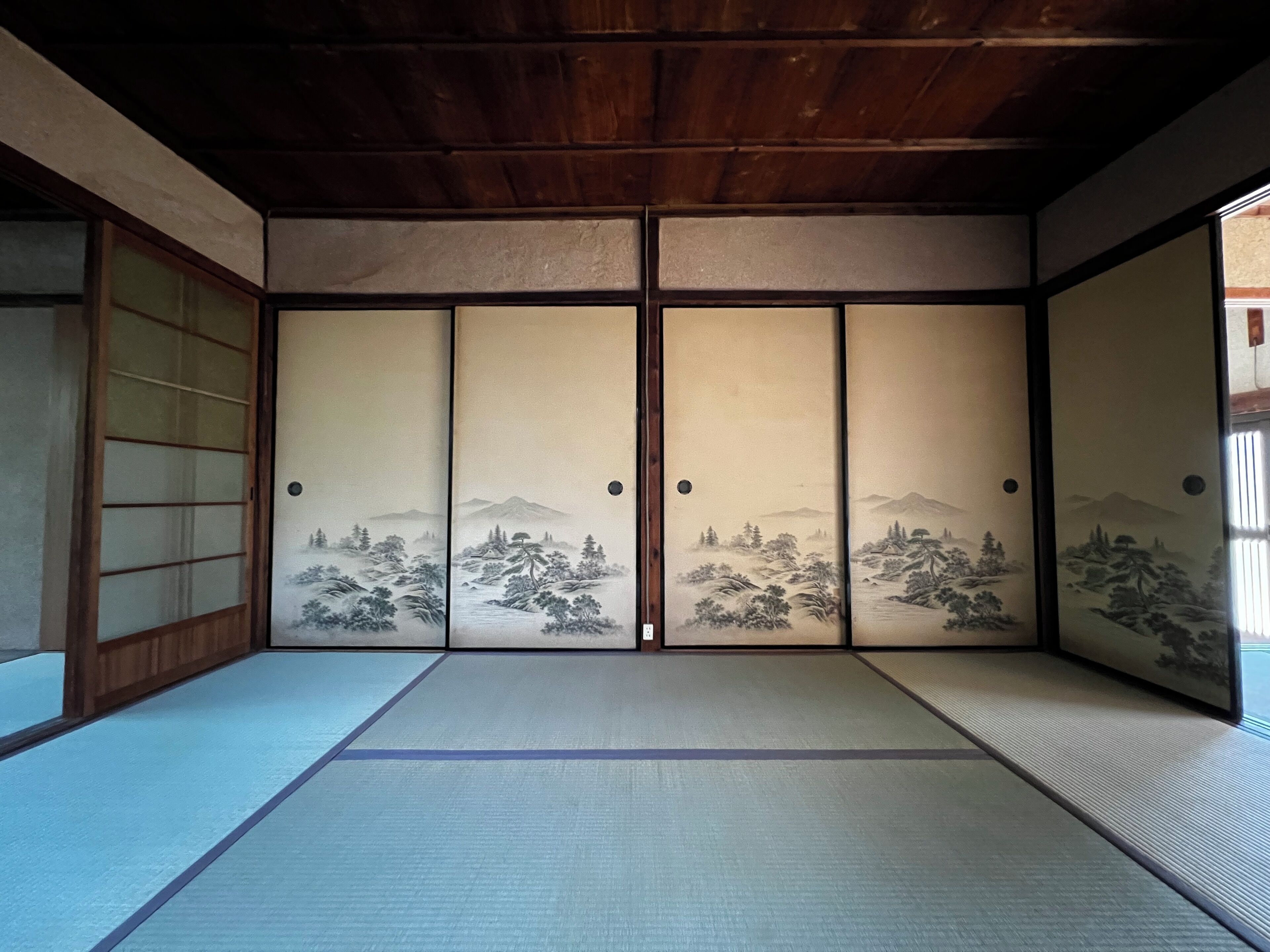 Una habitación japonesa tradicional con suelo de tatami, puertas correderas con pinturas de paisajes y techo de madera.