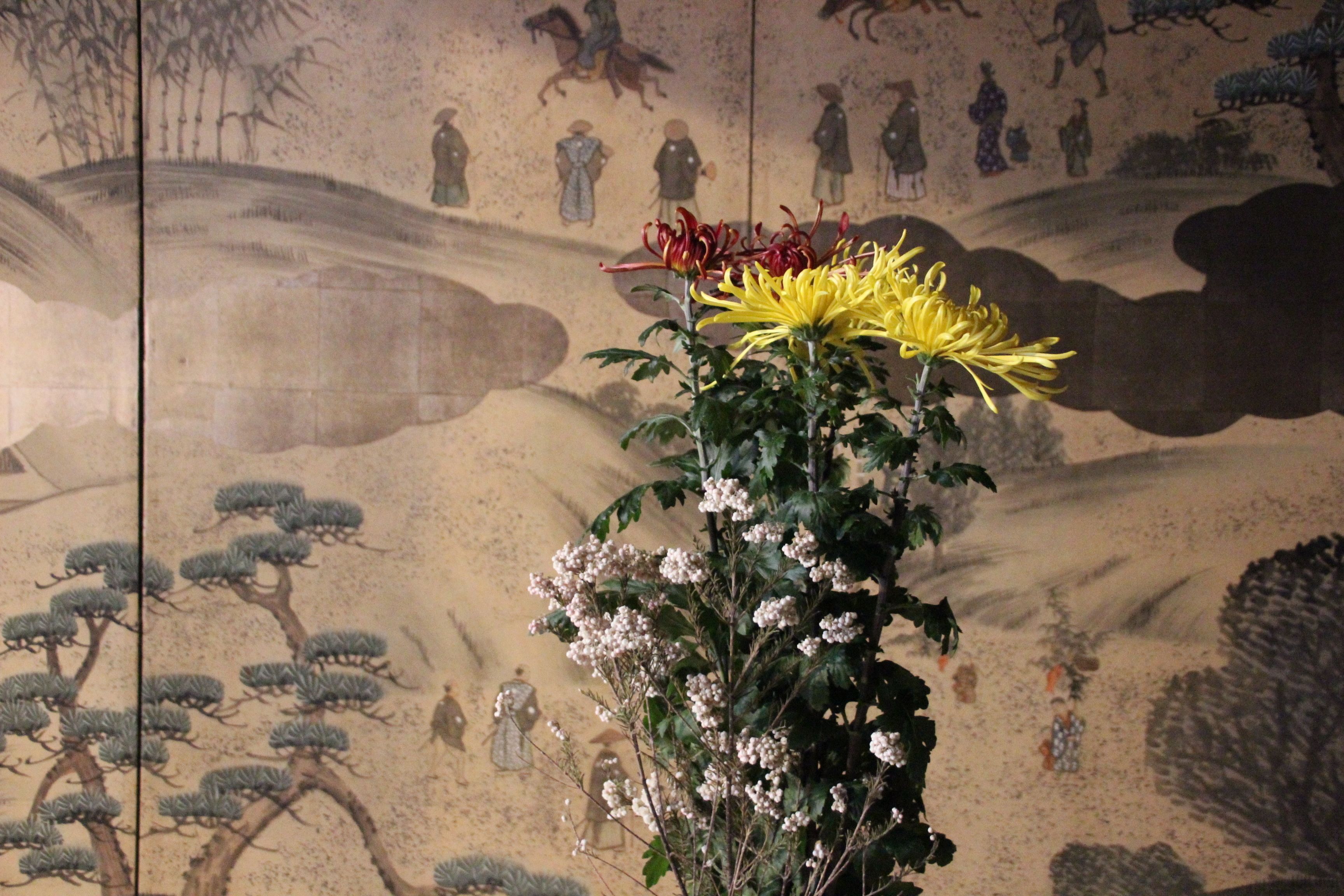 A traditional painting with figures and horses, a yellow flower, and white blossoms.