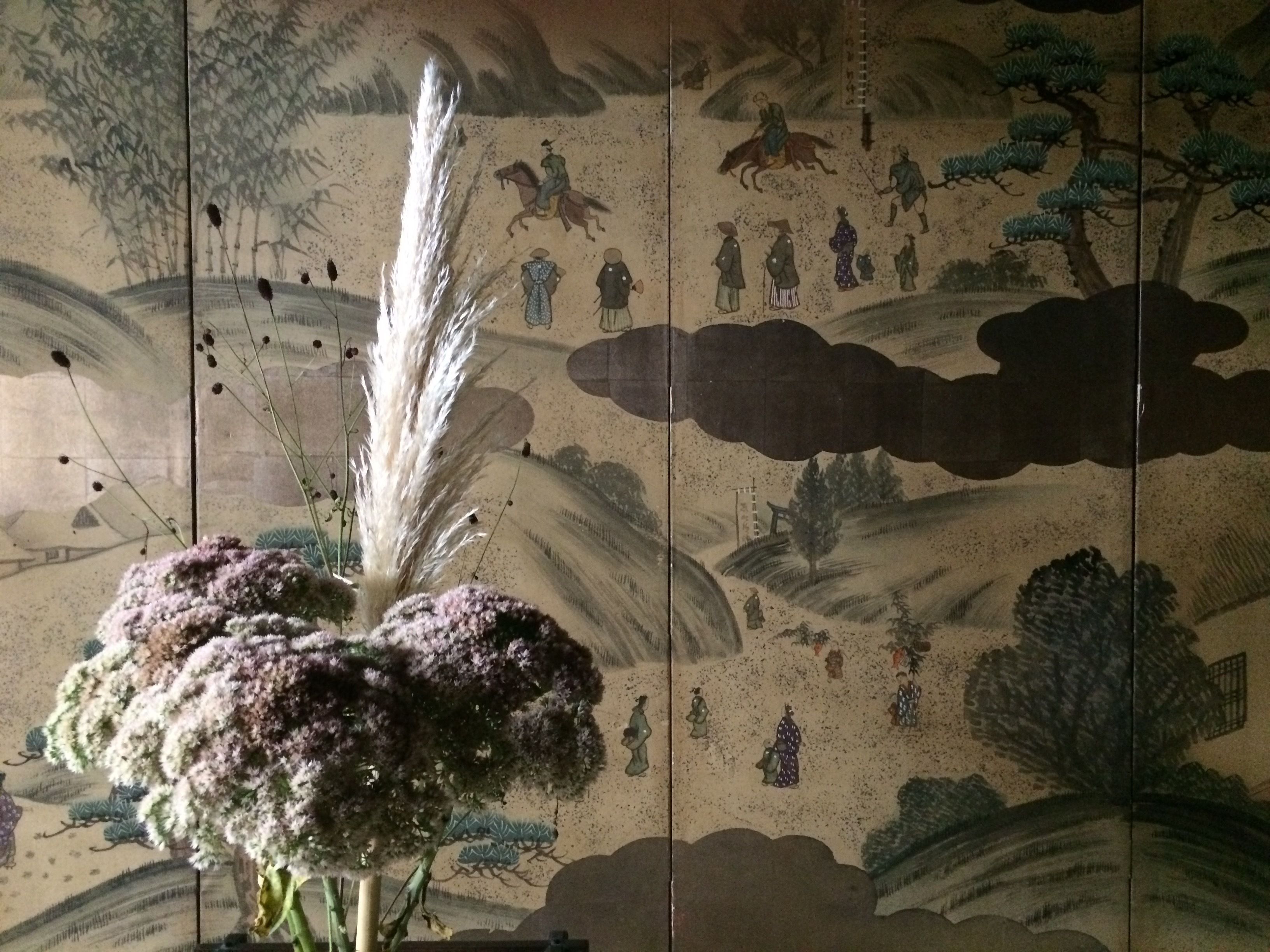 A room with a traditional Japanese-style mural on the wall, a large decorative plant in the foreground, and a partially visible traditional Japanese sliding door.
