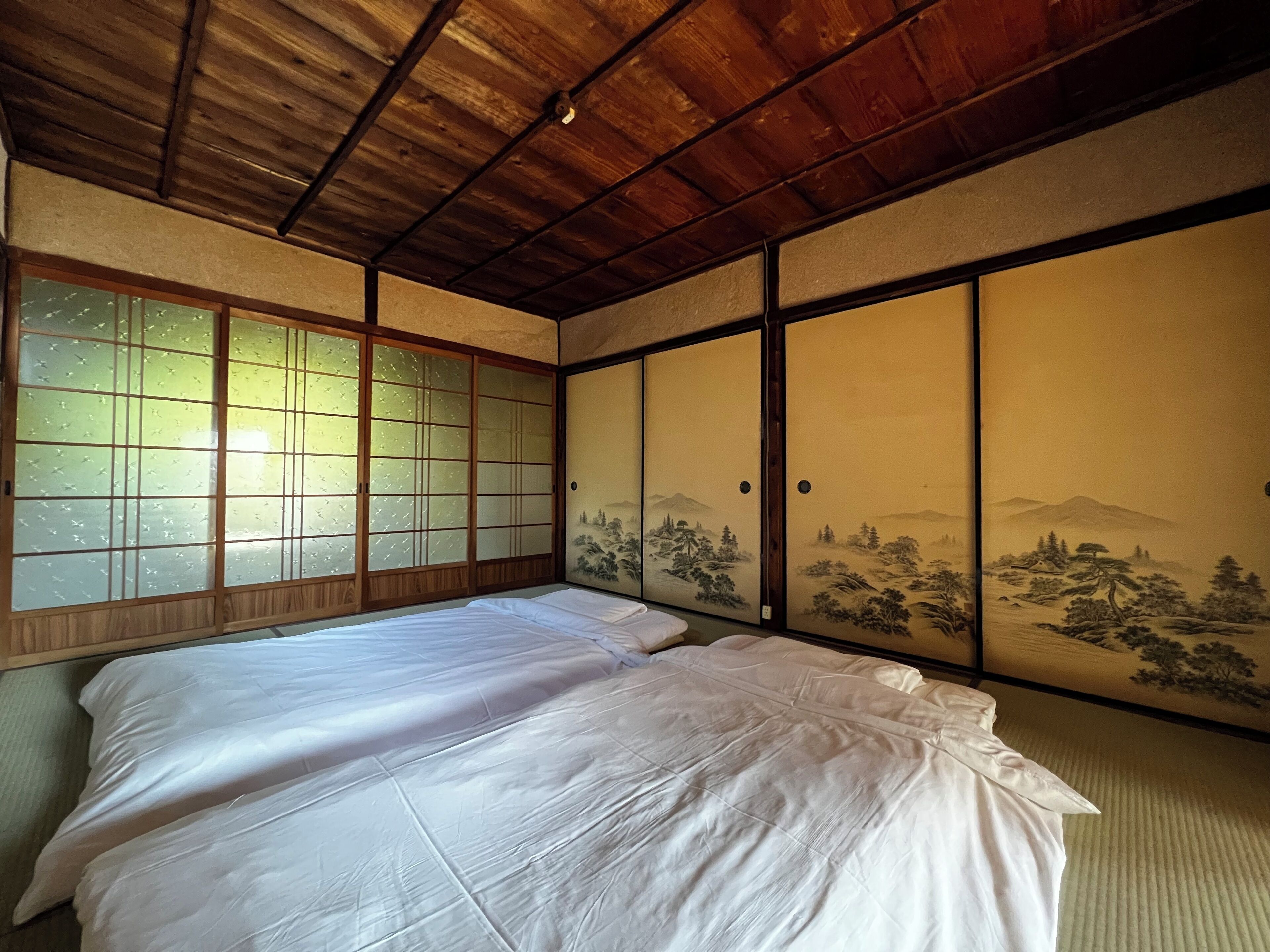 A traditional Japanese room with tatami flooring, a low wooden ceiling, sliding doors with landscape paintings, and a bed with white bedding.