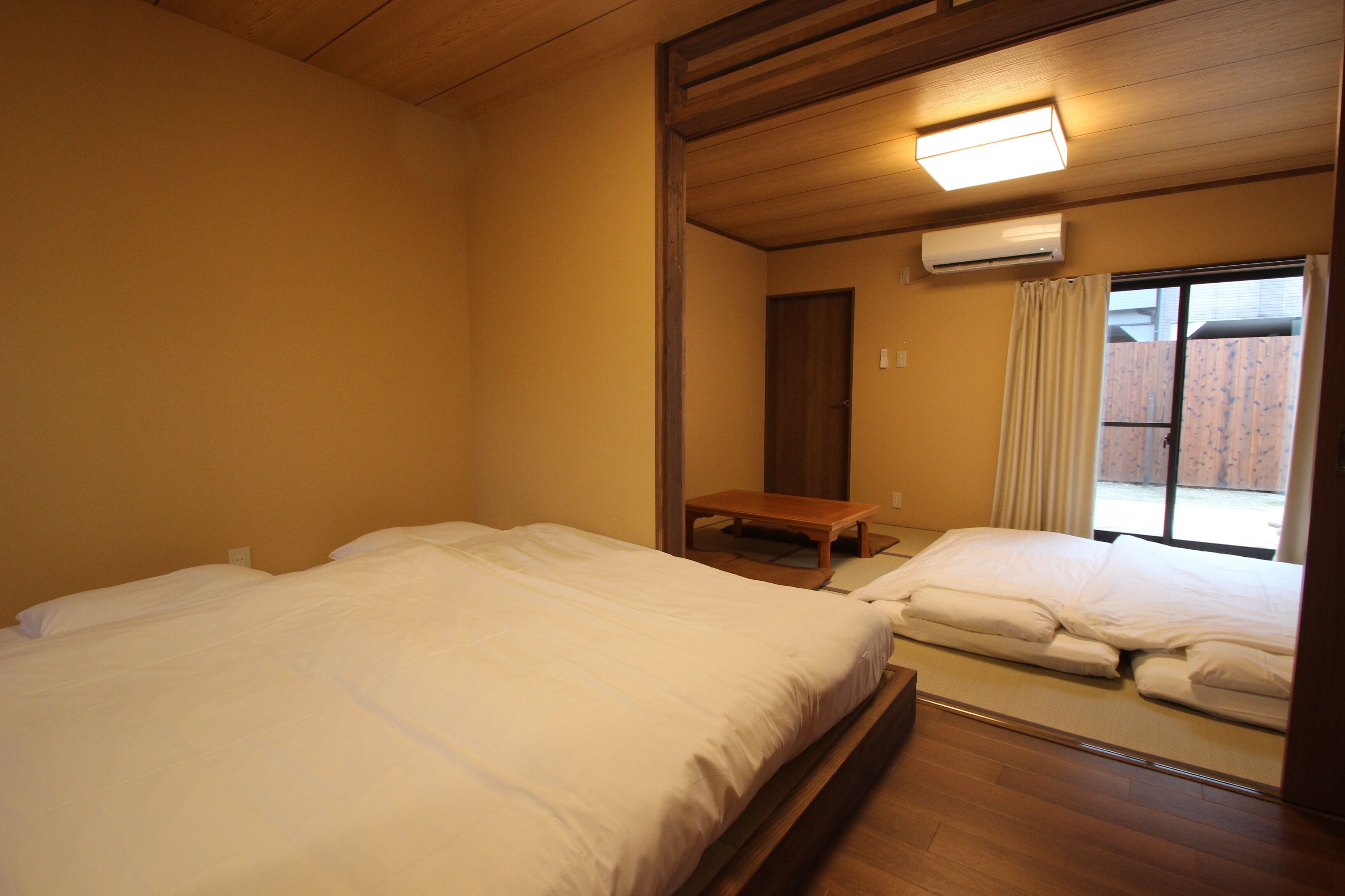 A room with two beds, a wooden bench, an air conditioning unit, and a window with curtains.